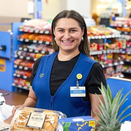 A smiling store associate in a blue vest standing at a checkout lane